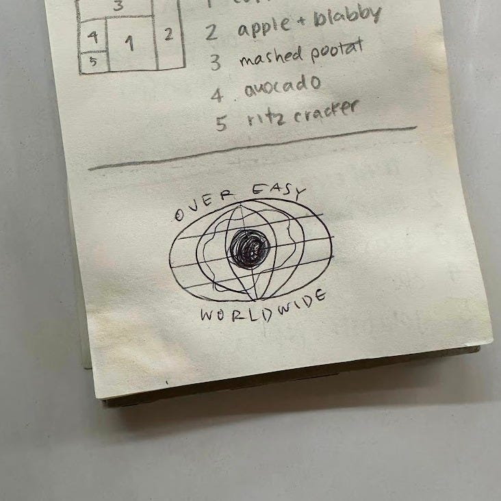 A photo of a small notebook with a ballpoint pen sketch of the logo idea: a retro-looking wide globe with a fried egg inside. Above the sketch there is another annotated sketch of our kid's lunch plan