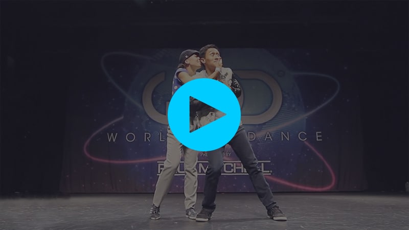 A still from a Youtube dance video. Keone and Mari Madrid are on the stage at World of Dance NY. They are mid-pose, with Mari holding Keone's head from behind and kissing him. Keone looks stoked.