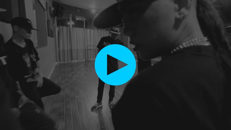 A still from a Youtube dance video by Parris Goebel. It's black and white. In the foreground, Parris' flat-brimmed profile is visible. She's just walked into a room of men (they are dancers lol), and it's the moment where Parris realizes it's just her. Don't worry, it ends well.