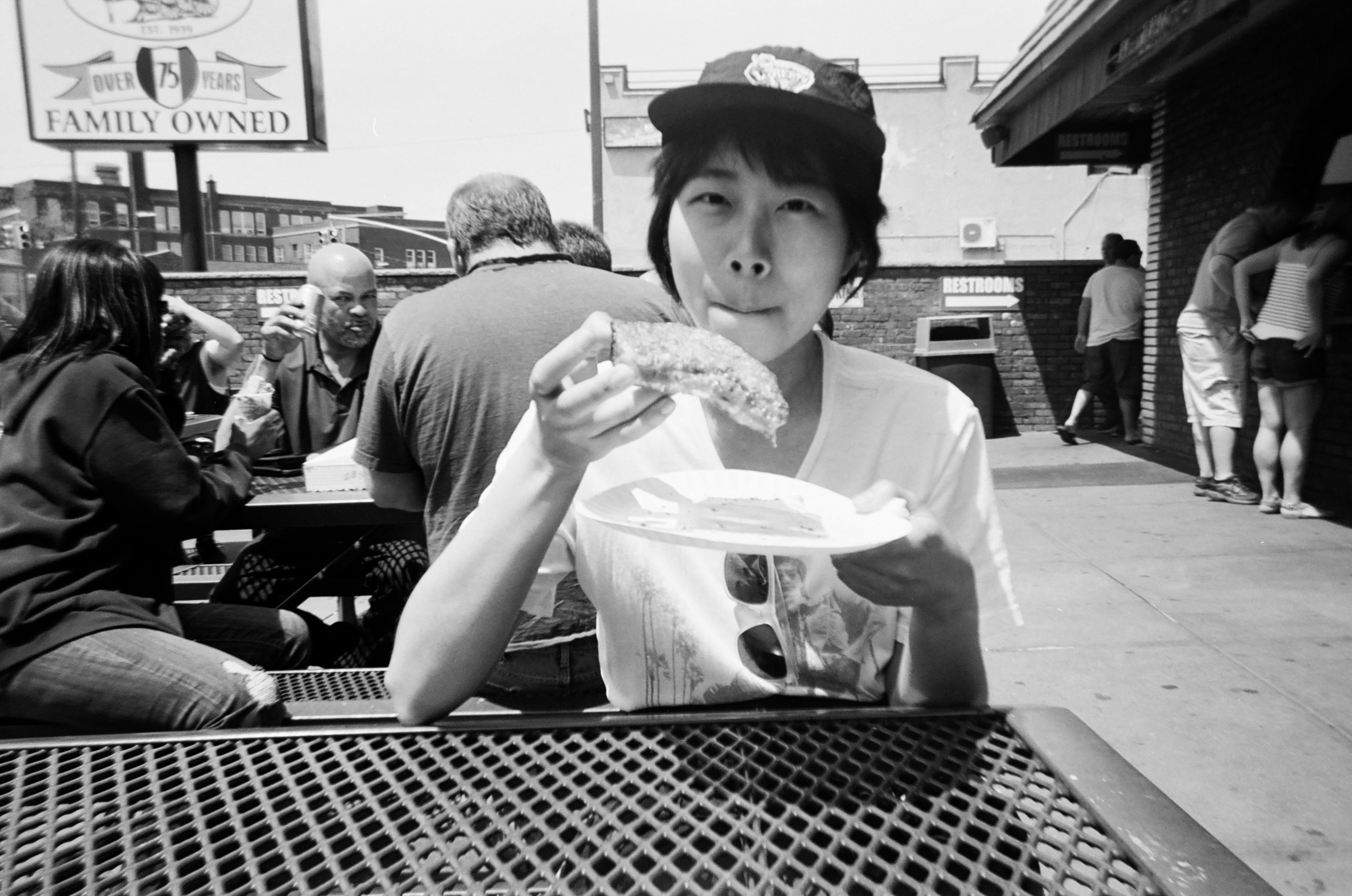 A black and white photo of me, sitting at an outside eating area, eating a thick rectangular slice of pizza off a paper plate. I’m wearing a vintage NY Liberty cap on top of my short hair