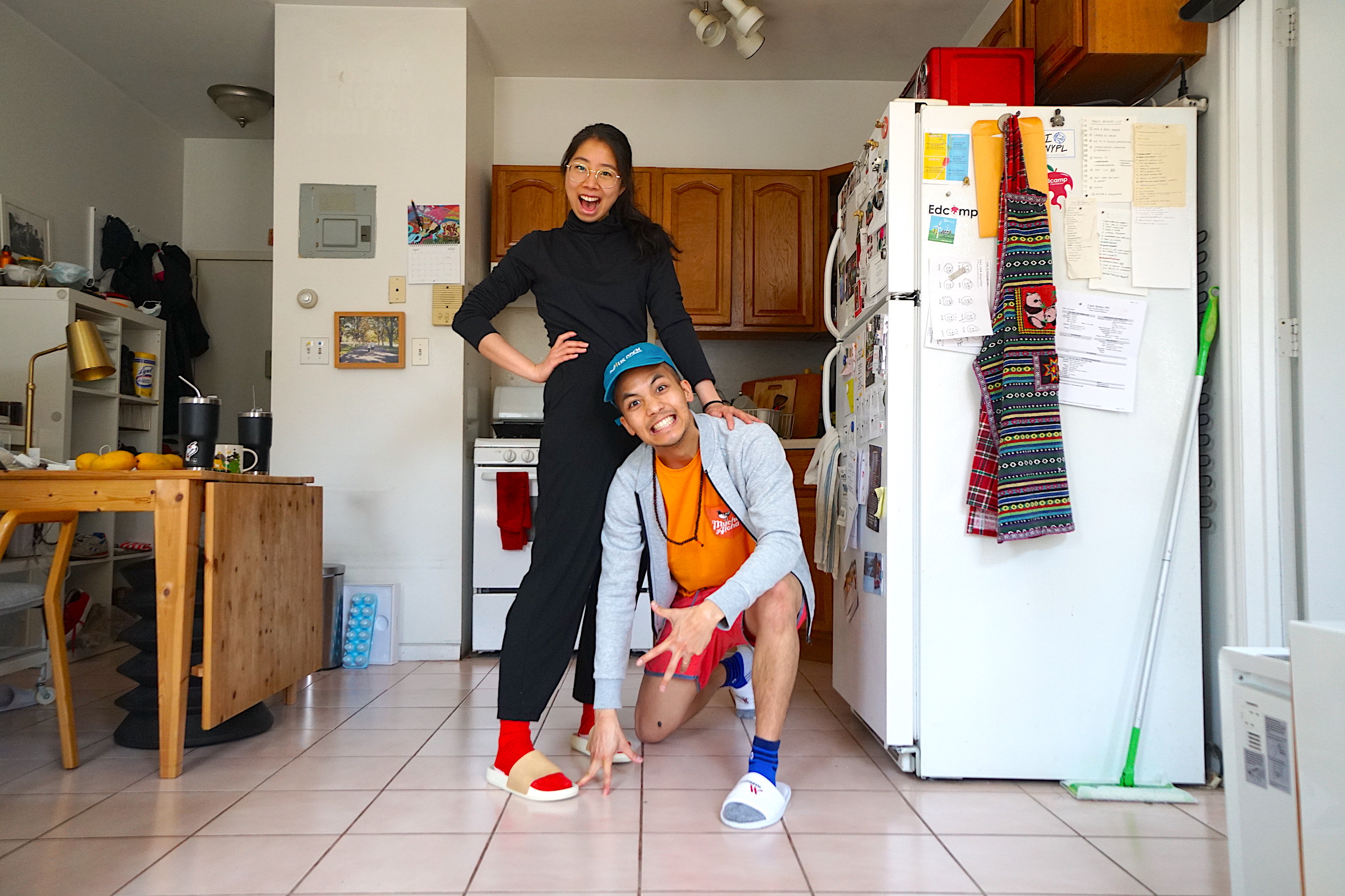 A color photo of me and Kevin posing in our apartment. We’re dressed like we might go outside, except we’re both wearing indoor slippers. Kevin is kneeling, and I’m standing up straight with my hand on his shoulder.