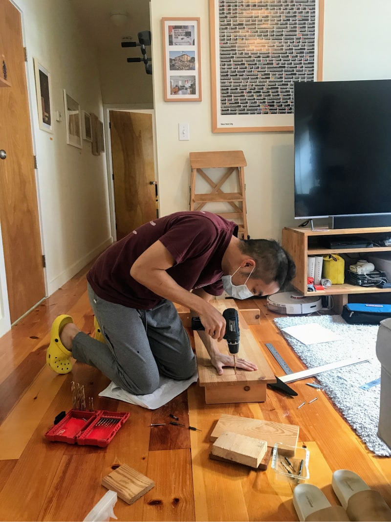 Kevin making his shelf. He is kneeling on the living room floor and drilling a pilot hole into the top plank and one of the legs. 