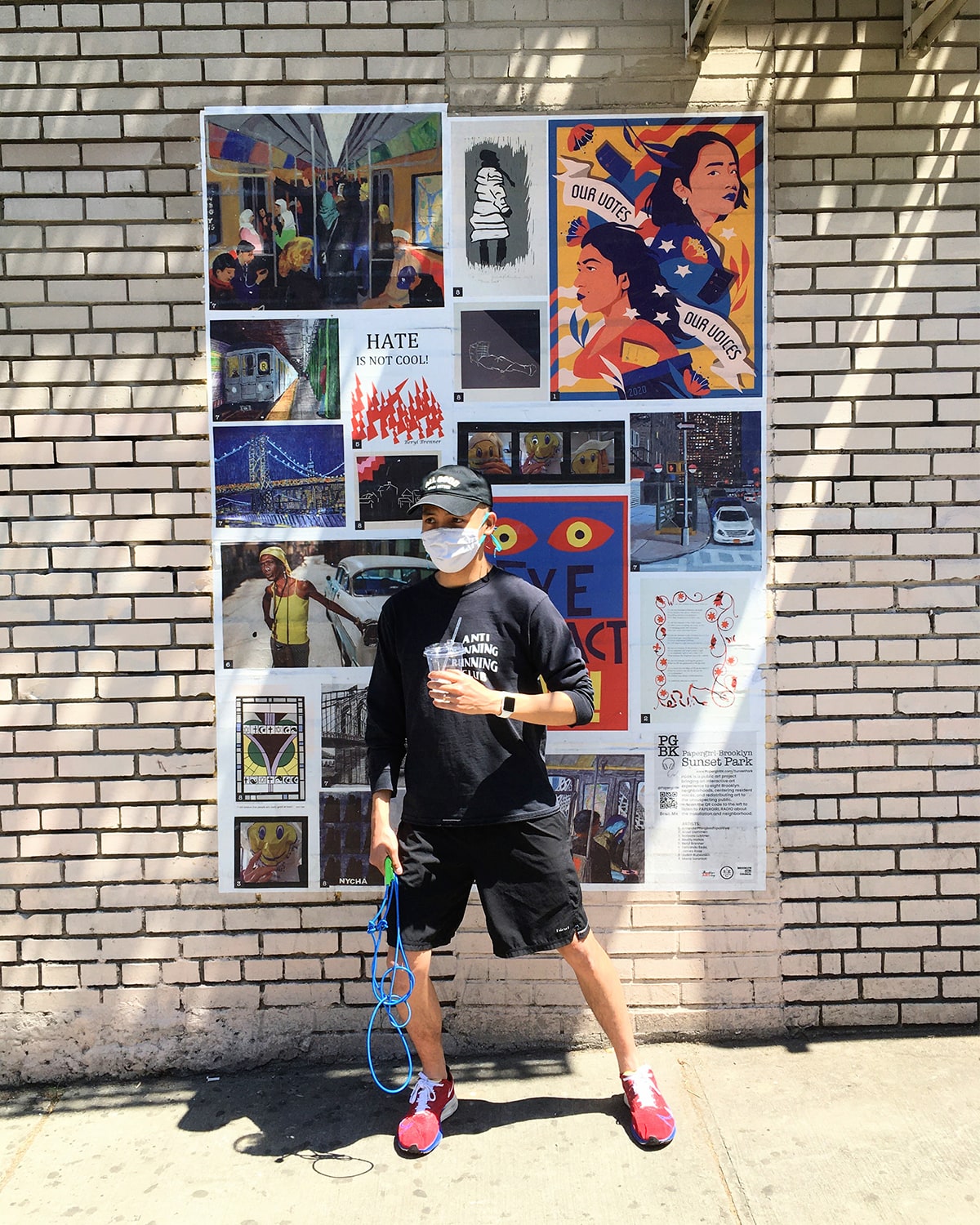 A photo of Kevin standing against a wall around the corner from Yafa. He’s wearing a baseball cap, a facemask, a “Anti Running Running Club” long sleeve, some black shorts, and a pair of red and blue trailrunning shoes. A collection of community artwork is wheatpasted on the wall behind him.