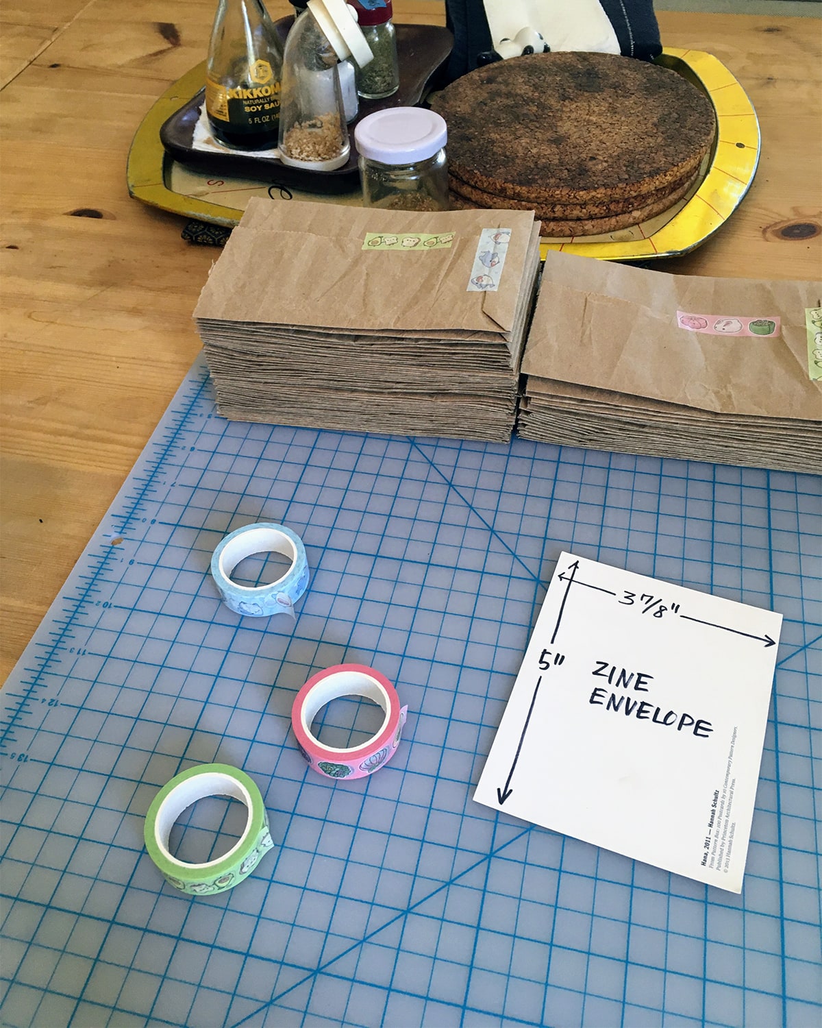 A photo of cutting mat on the kitchen table. On the mat there are two stacks of envelopes made from old recycled paper packing materials. The envelopes are held together with washi tape from KyariKreations on Etsy, a few rolls of which are also on the mat. There is also a zine-shaped pattern made from an old postcard, used as a guide for making the envelopes, which are for sending the zines.