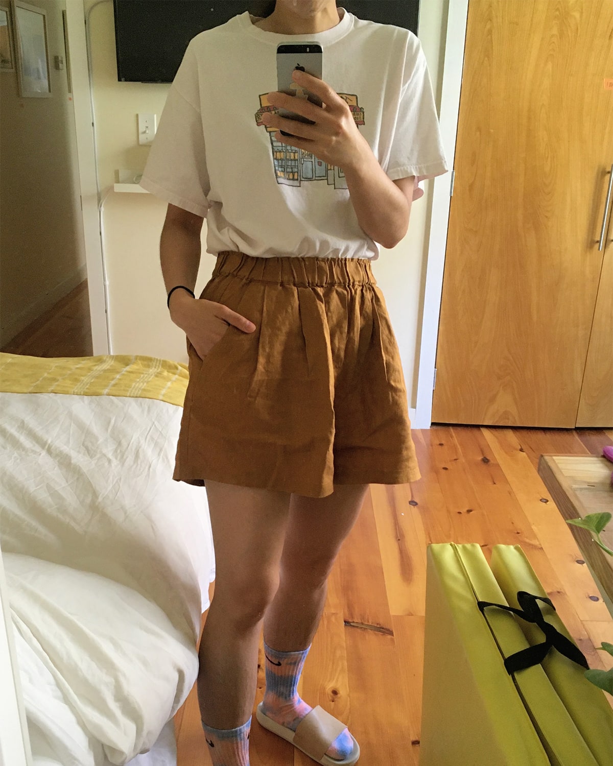 A photo of me standing in front of our mirror. I’m wearing a tee shirt I got from Yafa cafe and a new pair of shorts, also bought on Etsy. The shorts are a dark brownish mustard color, and they come up the waist pretty high. They have POCKETS (in all caps) as you can see I have my hand in one.